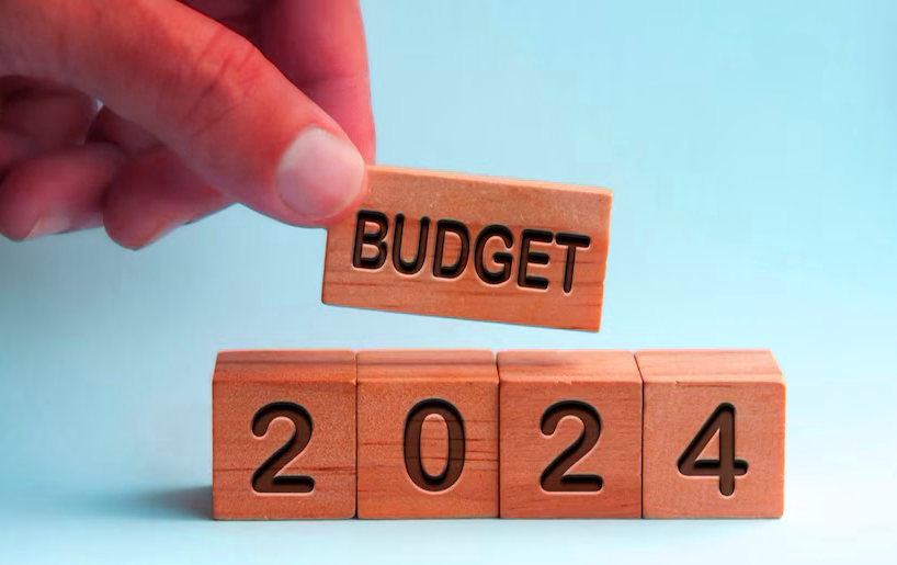 The image shows a hand holding a rectangular small board on which the Budget name is embedded, placed on the other Rectangular board on which the year 2024 is mentioned.
